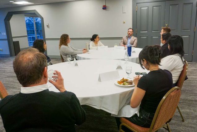 Photo: A group of individuals, mainly students, sit around two white clothed round tables.