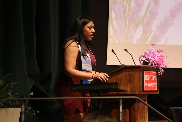 Photo: A woman with straight, dark hair speaking behind a podium that says "Boston University" wearing a lavender-colored sash and with a bunch of lavender next to the podium