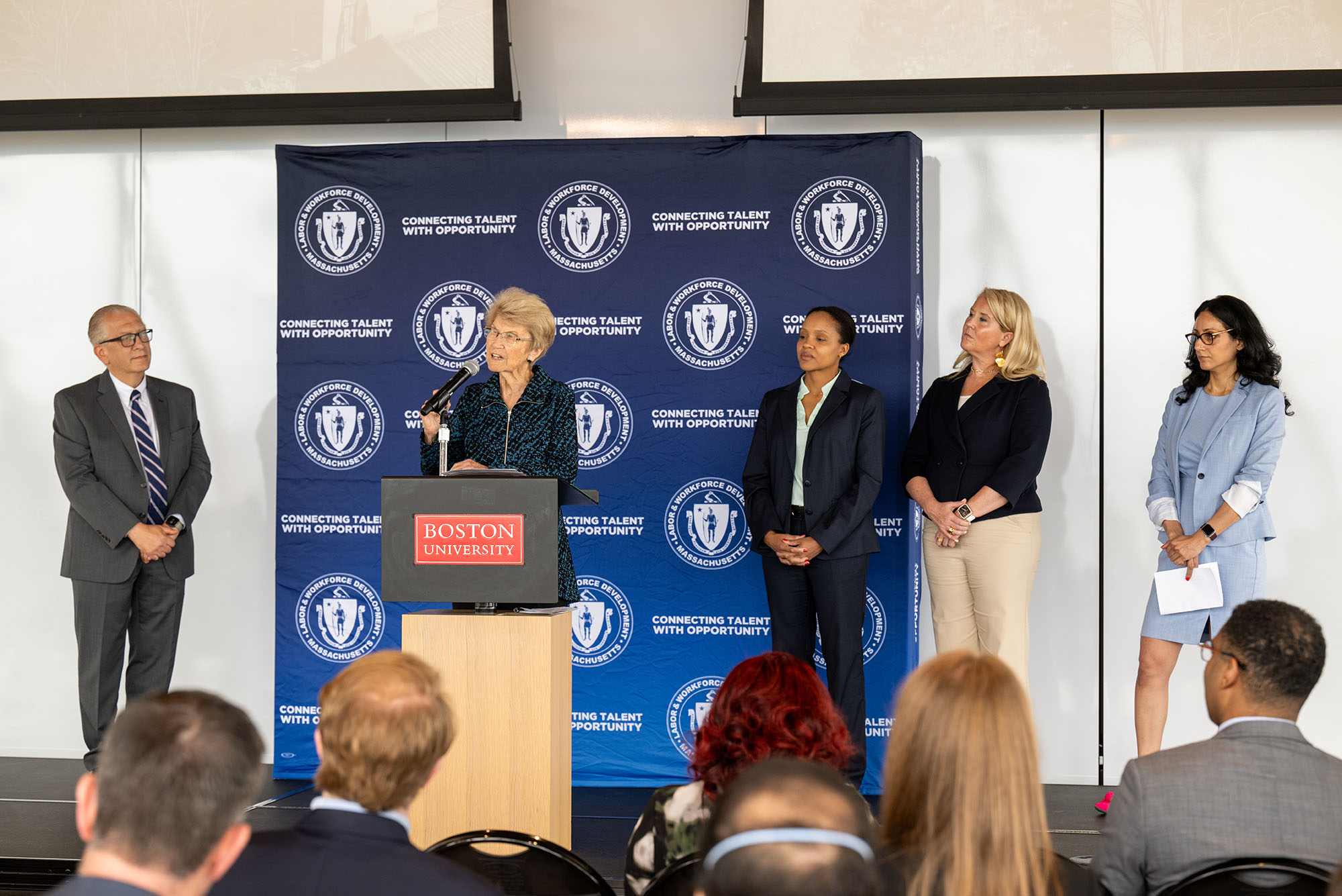 Photo: Five people standing in front of a back drop that says "Connecting Talent with Opportunity" on a stage with a podium that says "Boston University"