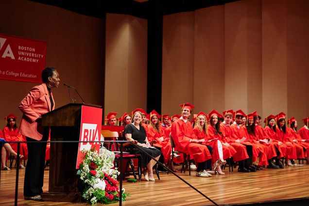 Photo: A wide shot of Dr. Melissa Gilliam at a podium in graduate regalia speaking to undergrads in red graduation regalia.