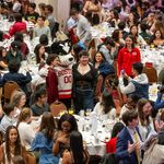 Photo: A large banquet hall filled with numerous round tables, each set with white tablecloths, plates, glasses, and cutlery. The room is crowded with people sitting at the tables and standing around. In the center of the image, a person in a Boston University mascot costume (Rhett) wearing a red and white jersey with "BOSTON 00" on it is posing for photos with attendees. Many people are taking pictures or looking at their phones. The atmosphere appears lively and social.