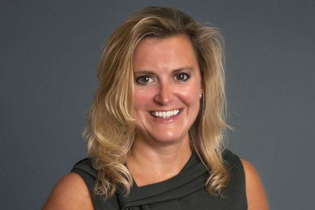 Photo: A white woman with blonde hair in front of a gray background smiles for portrait photo.