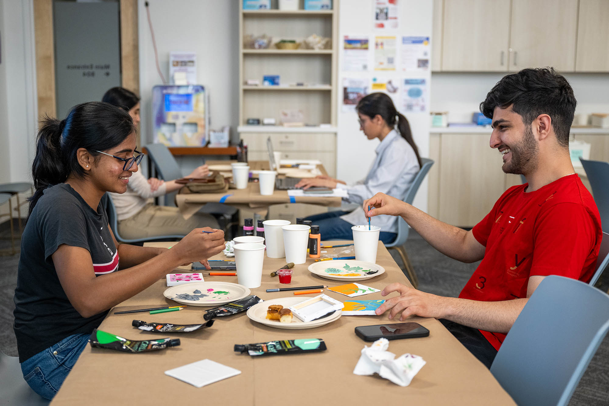 Photo: A woman on the left paints her canvas sitting across from a friend who is wearing a red shirt as he smiles at her.