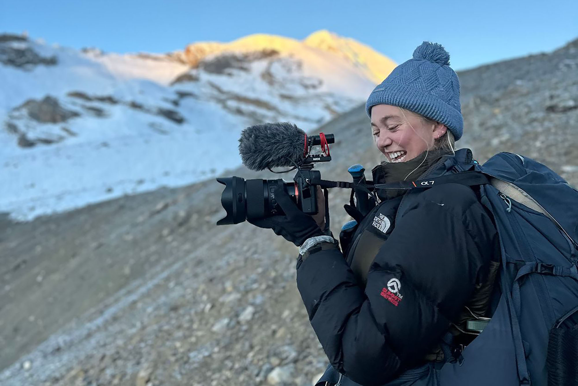 Photo: A blonde woman with a knitted cap and black thermal gear holding a black camera with snowy mountains in the background.
