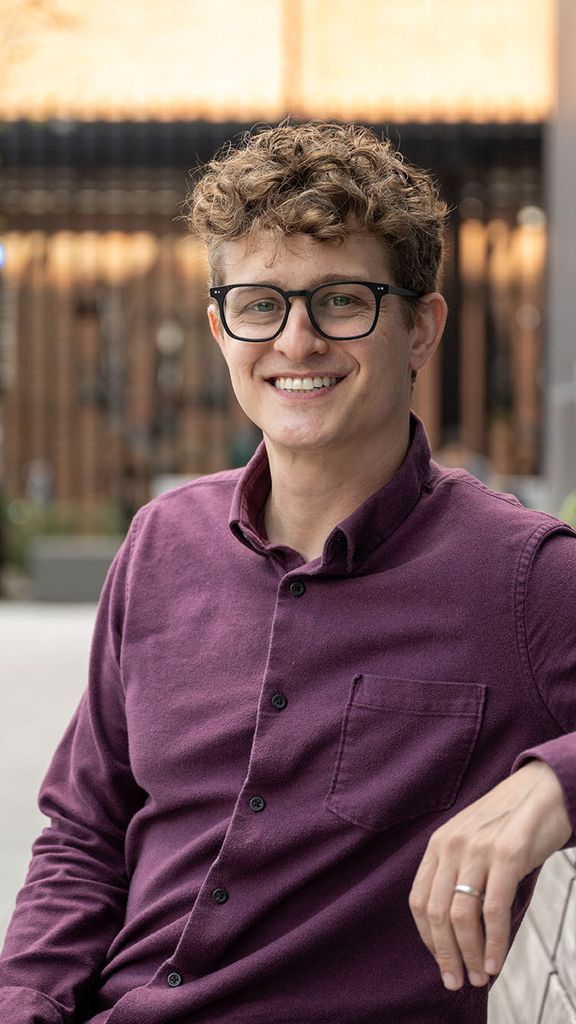 Photo: A young man wearing glasses poses for a portrait on a bench on a sunny day