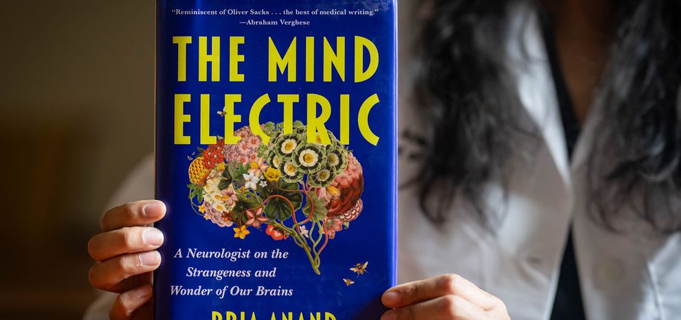 Photo: Pria Anand holding up her book "The Mind Electric," which features a vibrant blue cover and an illustration of the brain with flowers making up different parts