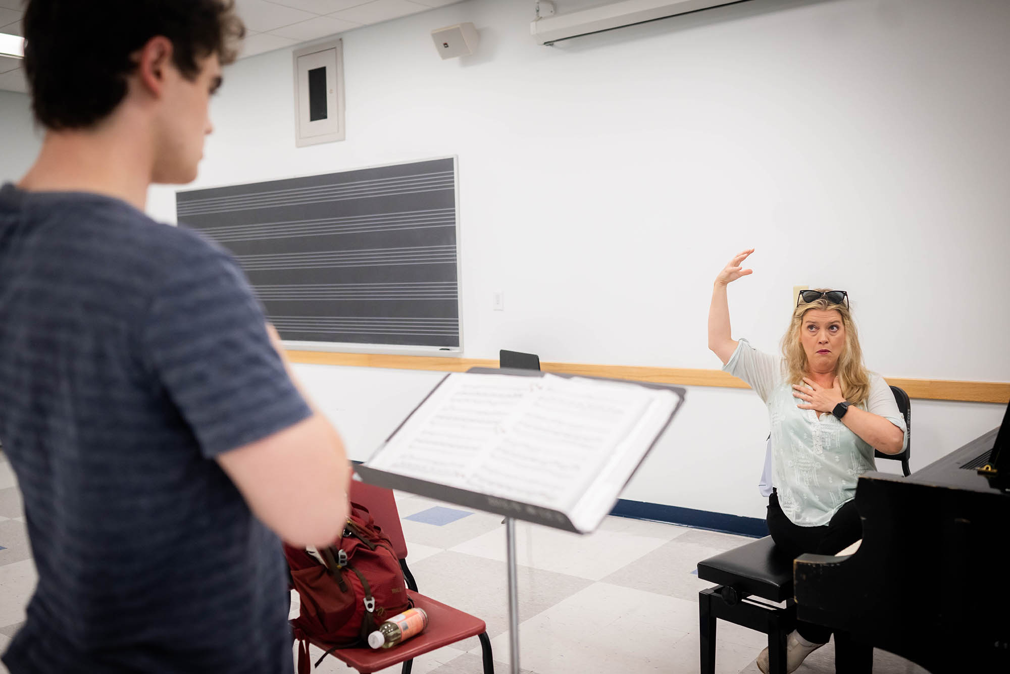 Photo: A young man in the foreground wearing a gray shirt receives feedback from his voice teacher, who sits across from him at the piano giving a vocal direction with her arms.