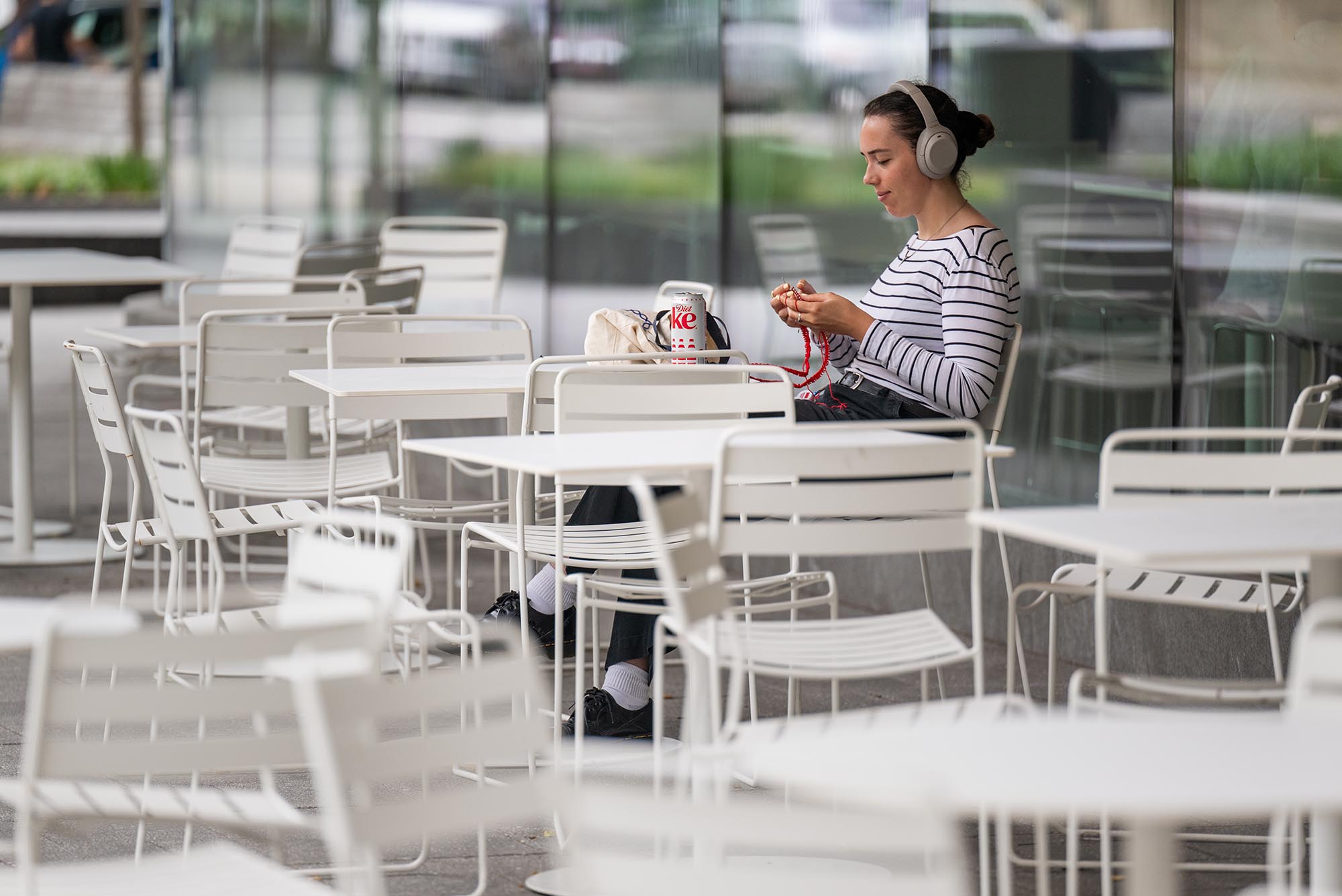 Photo: A young woman wearing headphones and a striped shirt knits red fabric outside.
