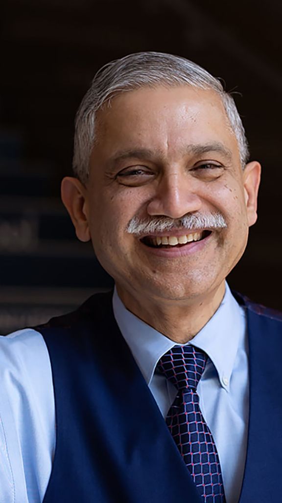 Photo: A man wearing a blue dress shirt and a navy blue vest with a tie leans on a stairwell and smiles for a portrait shot.