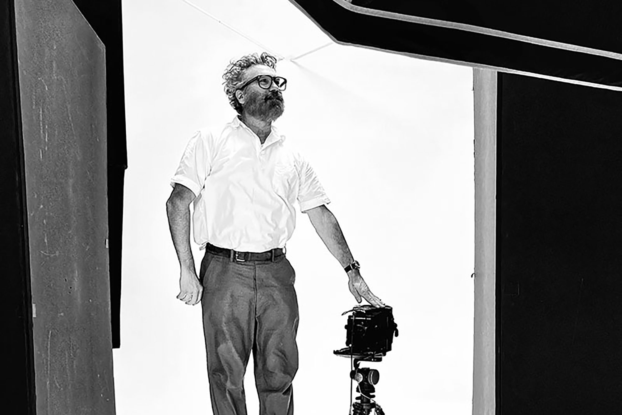 Photo: A black and white photo of a man in a white button up with dark slacks. He rest his hand on a vintage camera.