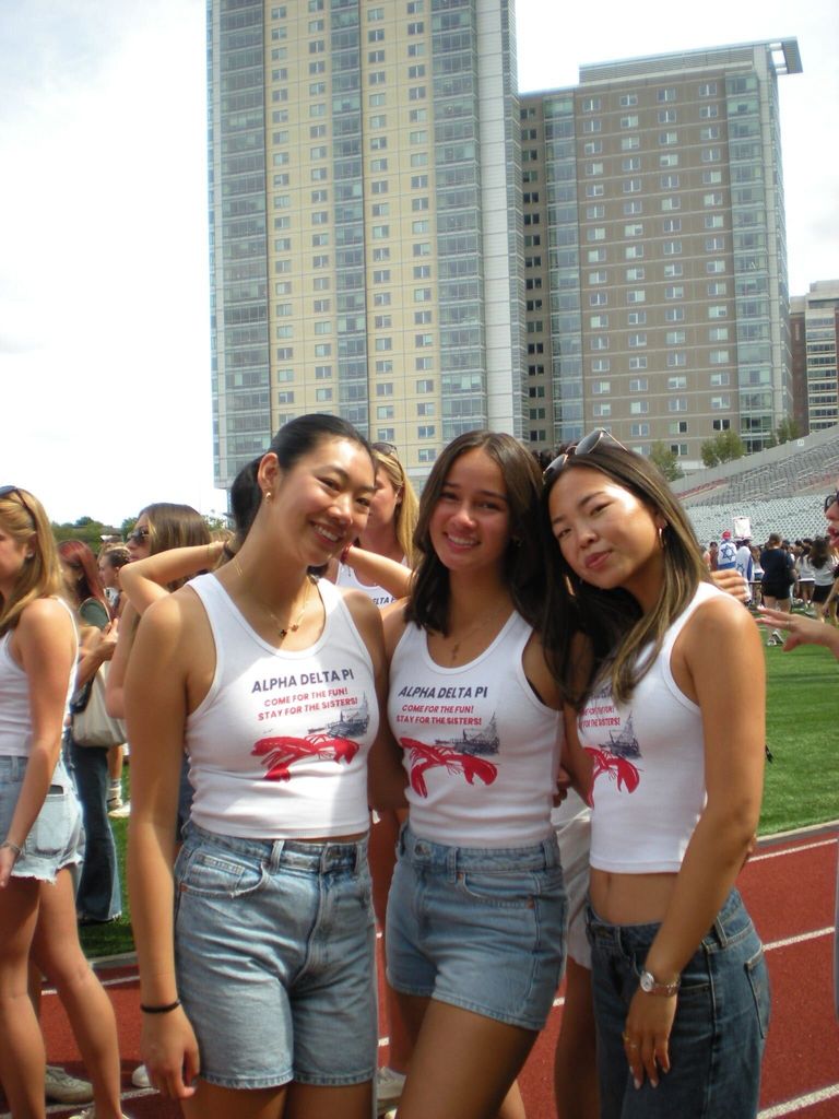 Three people stand together on a track field in front of tall apartment buildings, dressed in matching white Alpha Delta Pi tank tops with denim shorts, while others mingle in the background at what appears to be a lively outdoor event.