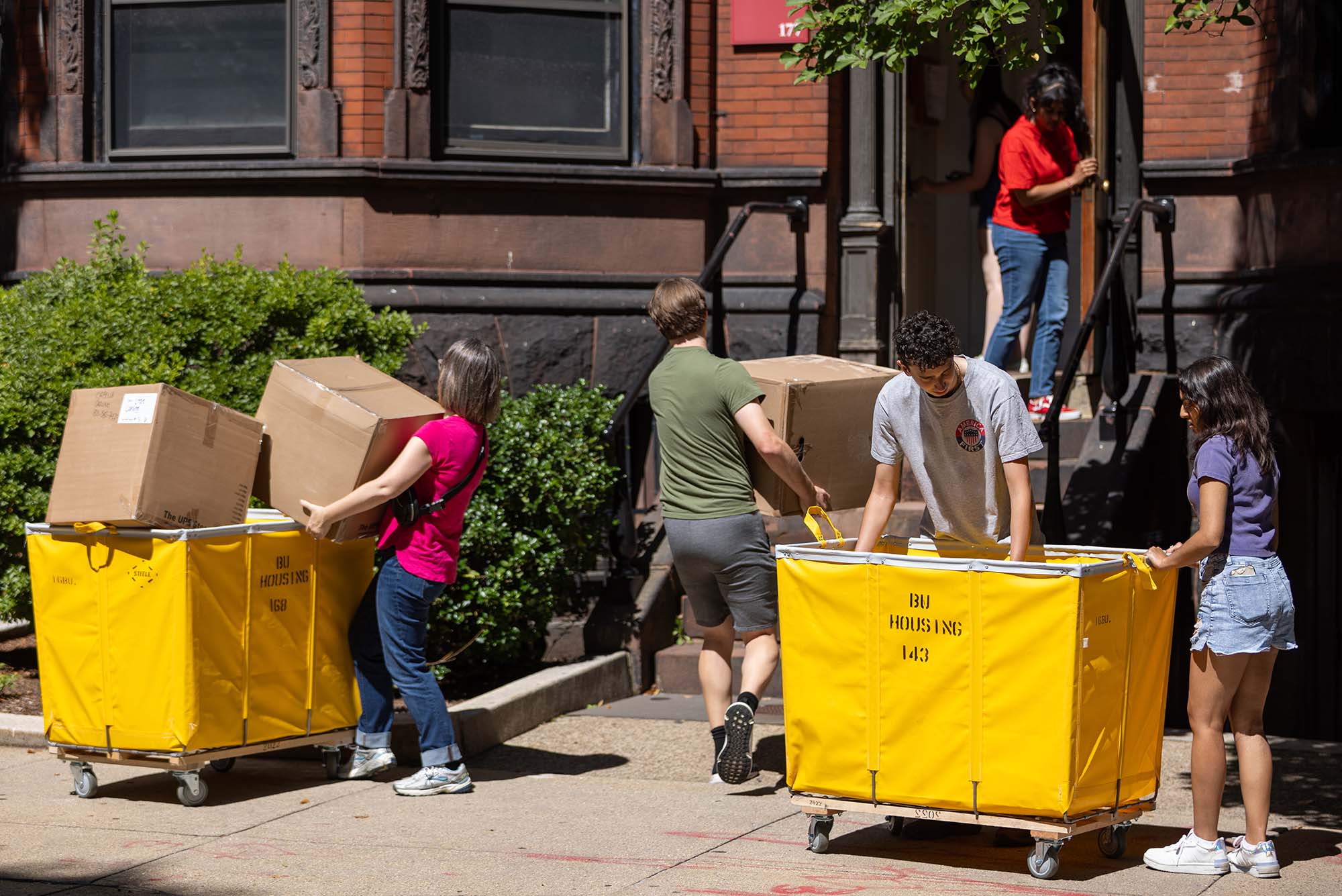 Photo: Various people in summer clothing lifts boxes out of yellow carts for BU move in.