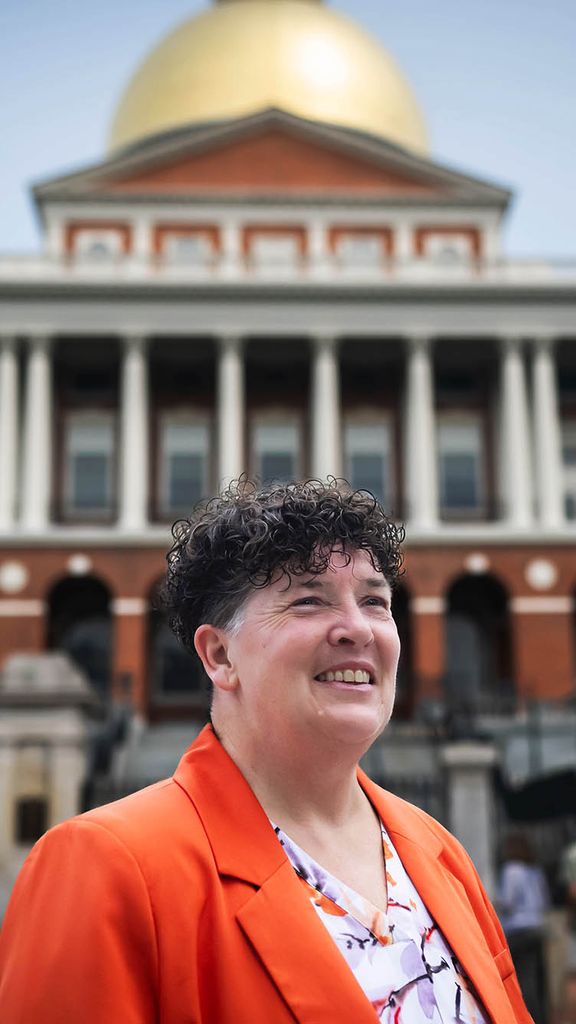 Photo: A white woman with short, tapered hair wearing a bright orange blazer poses in front of the Massachusetts State House.