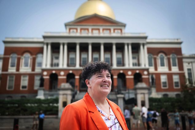 Photo: A white woman with short, tapered hair wearing a bright orange blazer poses in front of the Massachusetts State House.