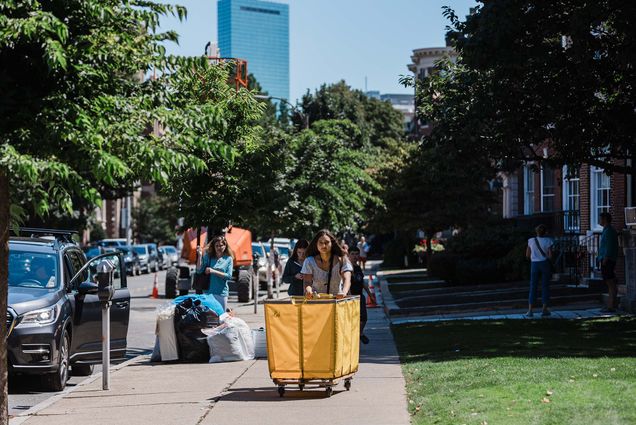 Photo: A BU freshman rolling a large, yellow bin during move-in with the Boston skyline behind her