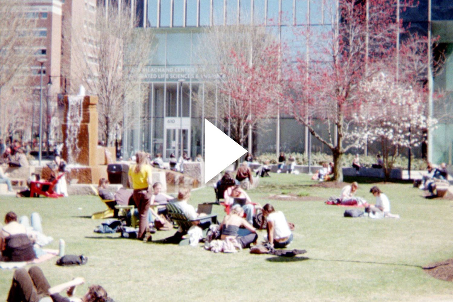Photo: College students lounging on BU's campus green space. A white play button is overlaid on top of the image