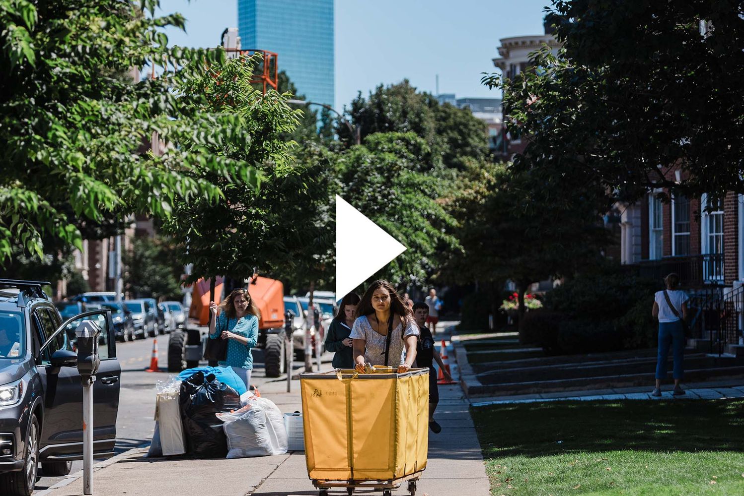 Photo: A BU freshman rolling a large, yellow bin during move-in with the Boston skyline behind her. There is a video play button over the image