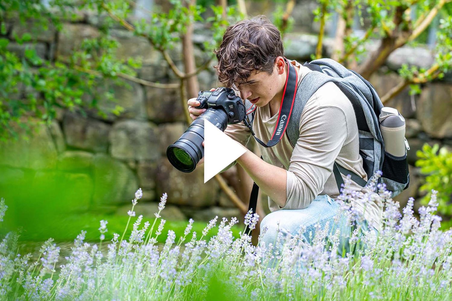 Photo: A person taking a photo, with a professional camera, of flowers in a field. There is a video play button over the image