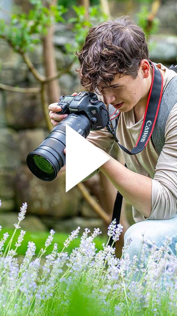 Photo: A person taking a photo, with a professional camera, of flowers in a field. There is a video play button over the image