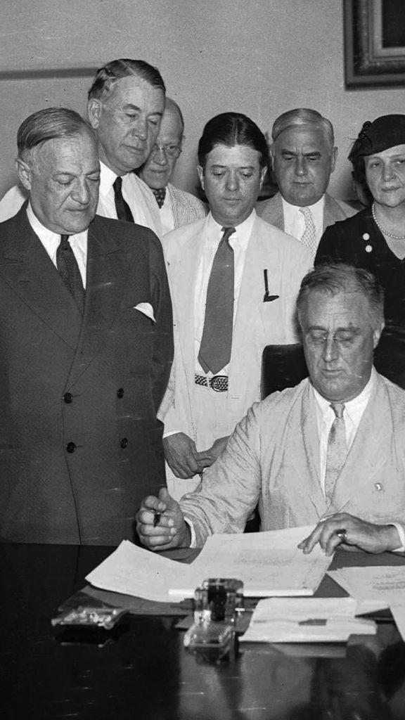 Photo: A black and white photo of various men standing around Franklin D. Roosevelt as he signs the Social Security Bill.