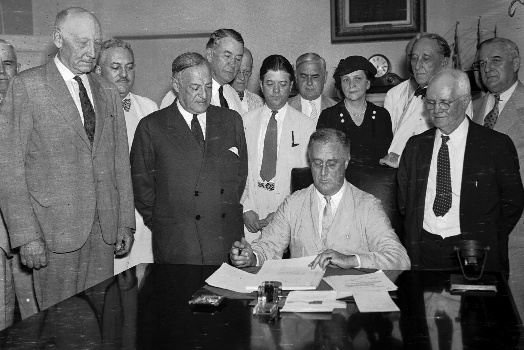 Photo: A black and white photo of various men standing around Franklin D. Roosevelt as he signs the Social Security Bill.