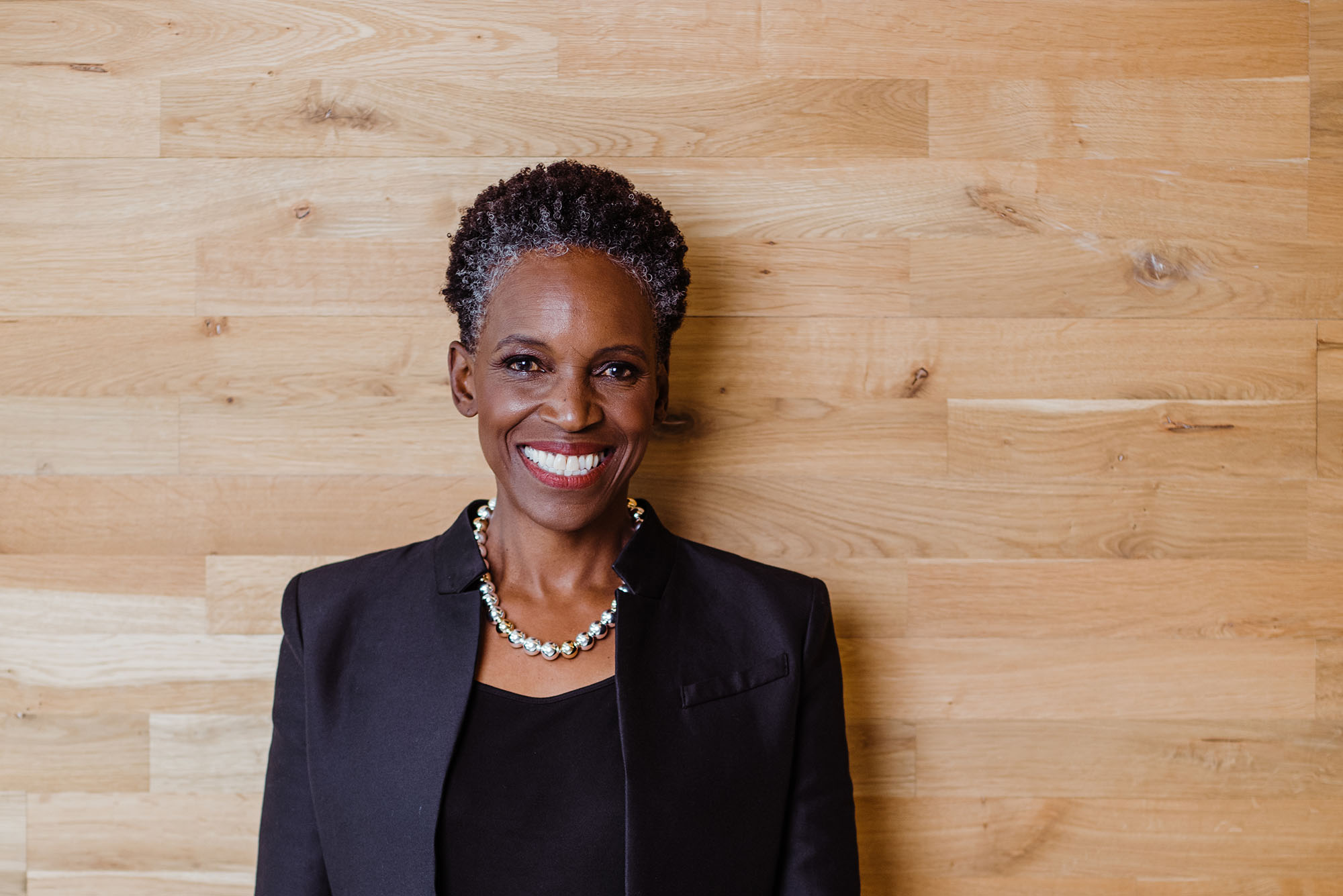Photo: A Black woman with a short grey and black afro and wearing a necklace of large, faux metal pearls, black blouse, and black blazer, smiles and poses in front of a large, wooden wall.