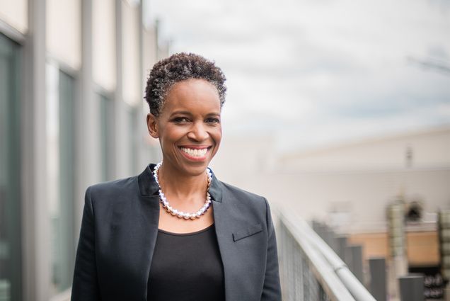 Photo: A black woman with short hair wearing a black blazer over a black shirt with a pearl necklace, smiling for a portrait in front of a blurred out cityscape