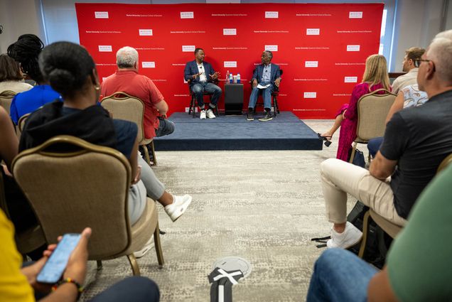 Photo: A room filed with students and at the center two people chat with a red background behind them.