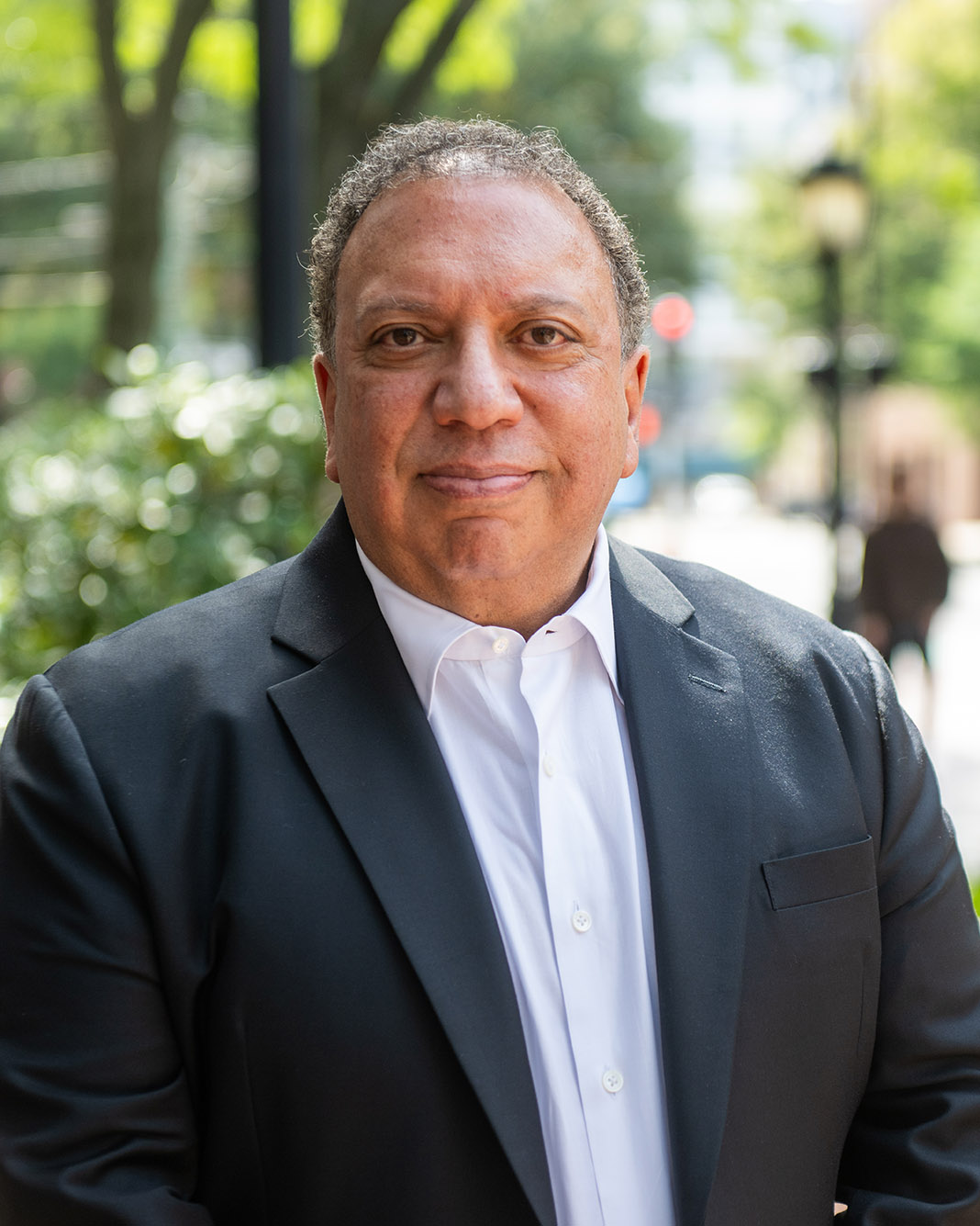 Photo of Ahmass Fakahany, who has short salt-and-pepper hair, in a black suit with a white shirt, smiling, as he stands on a sidewalk on a sunny day. In the blurred background greenery and passersby's can be seen.