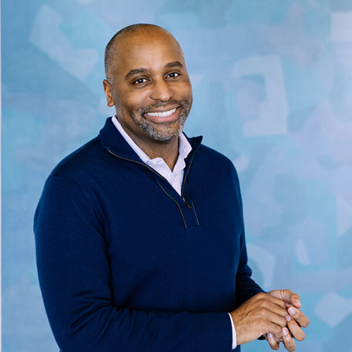 Portrait of Harvey Young, a middle-aged Black man. He wears a blue quarter zip sweater and smiles. He smiles and poses in front of a light blue background.