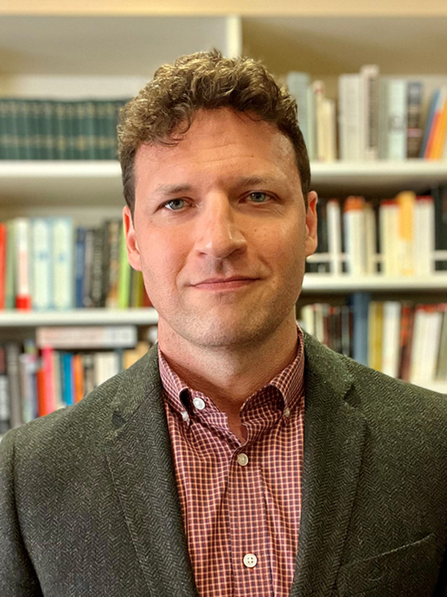 Photo: A man with curly blonde hair posing for a headshot in front of many bookshelves