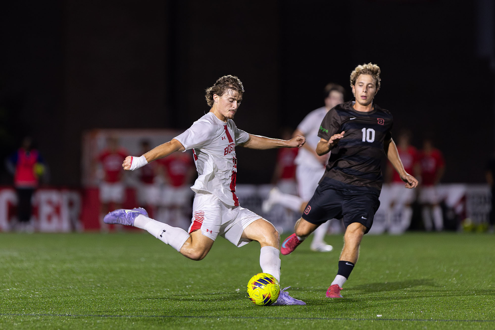 Photo: A BU Men's soccer player in a white uniform with red trim kicking the ball down the field