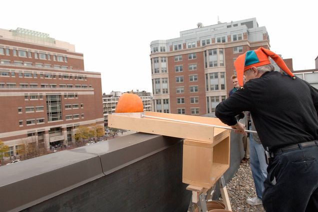 Photo: An early 2000s photo of a white man in a jester hat pushing a pumpkin off of a roof for a physics class.