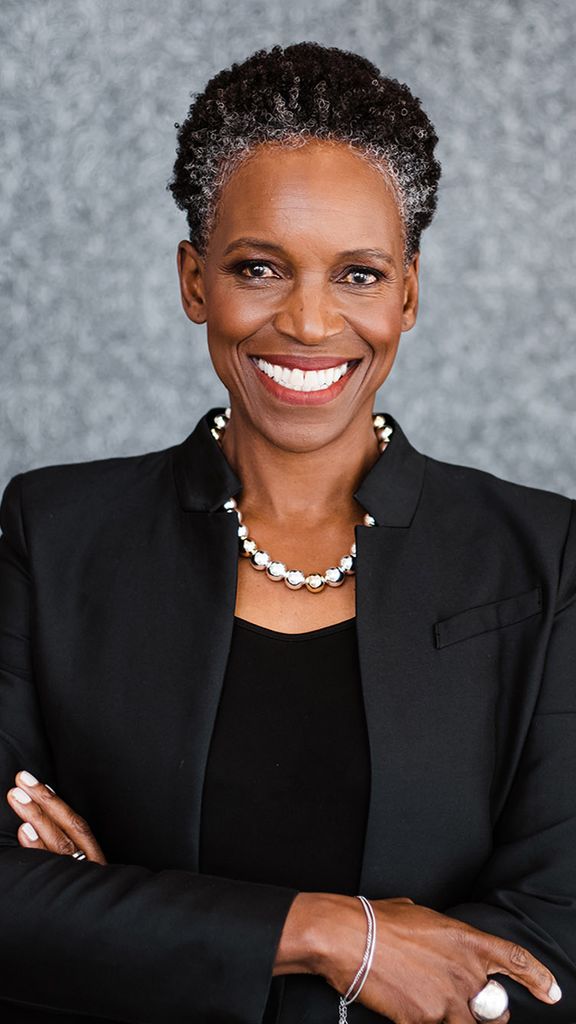 Photo: A Black woman with a short grey and black afro and wearing a necklace of large, faux metal pearls, black blouse, and black blazer, smiles and poses with arms crossed in front of a grey backdrop.