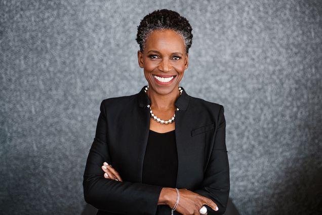 Photo: A Black woman with a short grey and black afro and wearing a necklace of large, faux metal pearls, black blouse, and black blazer, smiles and poses with arms crossed in front of a grey backdrop.