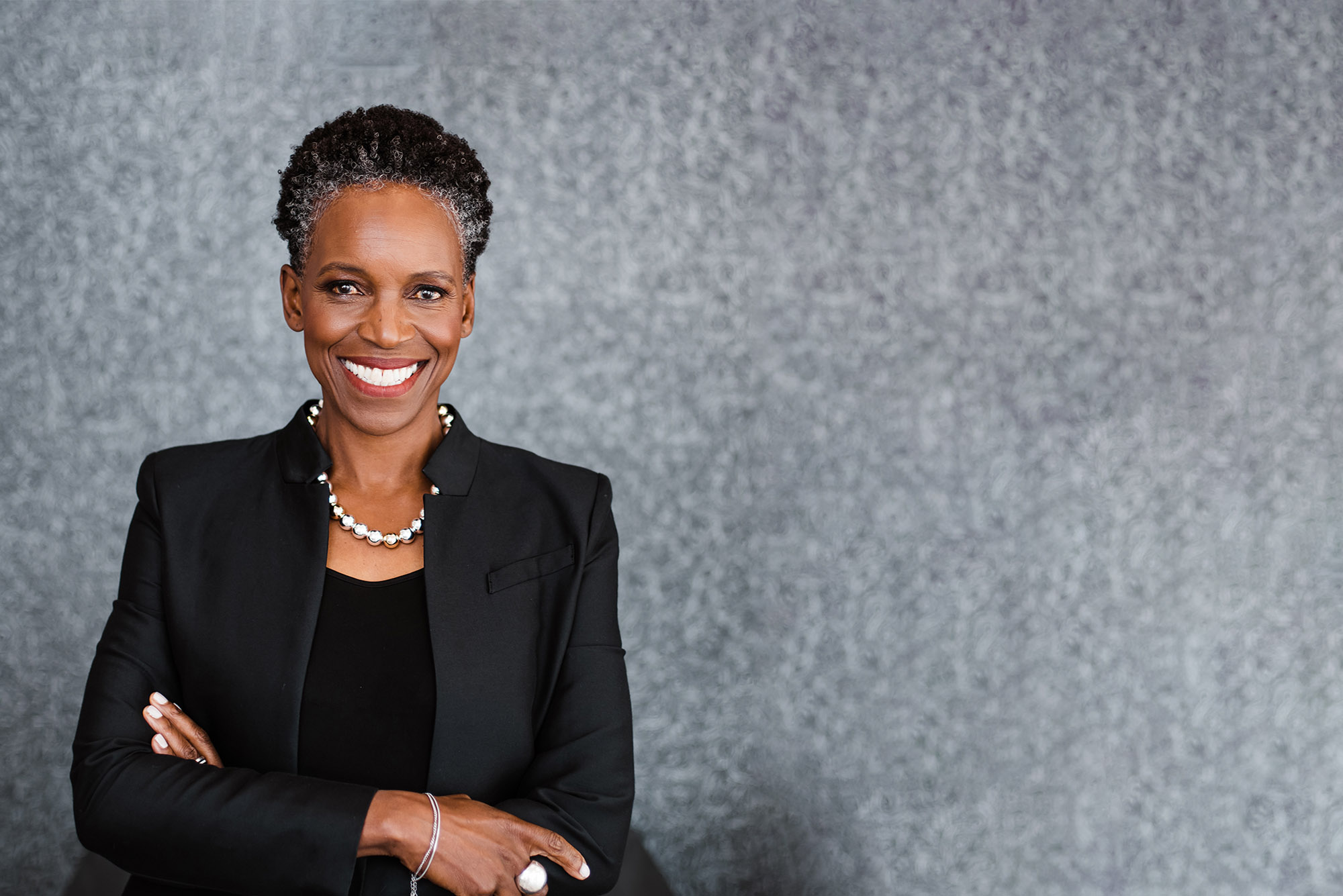 Photo: A Black woman with a short grey and black afro and wearing a necklace of large, faux metal pearls, black blouse, and black blazer, smiles and poses with arms crossed in front of a grey backdrop.