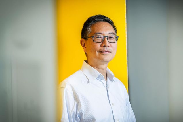 Photo: A portrait of Christopher Chen, an Asian man wearing a button up shirt posing against a gold background. He smiles for the photo.