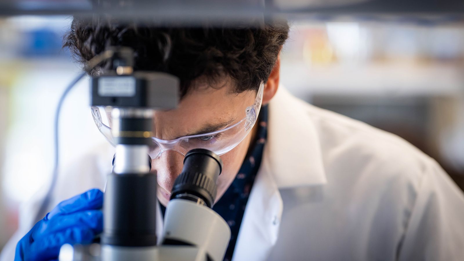 Photo: Miguel Jimenez, an engineer at BU, looking into a microscope while wearing a lab coat and safety glasses.