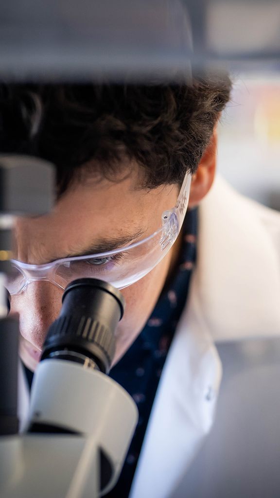 Photo: Miguel Jimenez, an engineer at BU, looking into a microscope while wearing a lab coat and safety glasses.