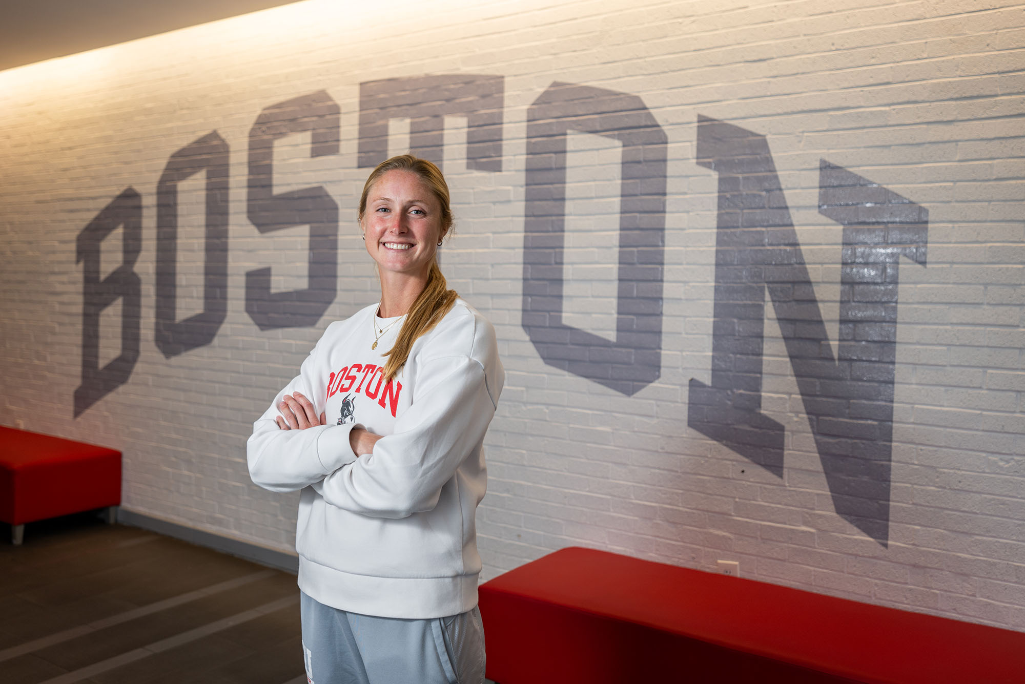 Photo: A white woman with blonde hair poses with her arms crossed in front of a brick wall with BOSTON painted in gray.
