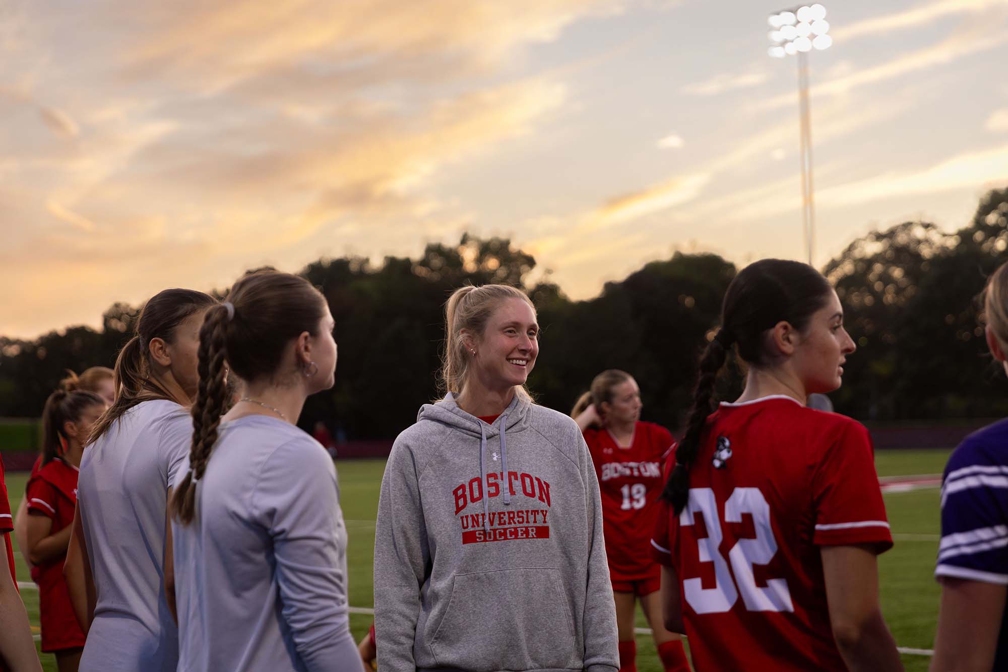 Photo: A white woman with blonde hair stands amongst players on a soccer field.