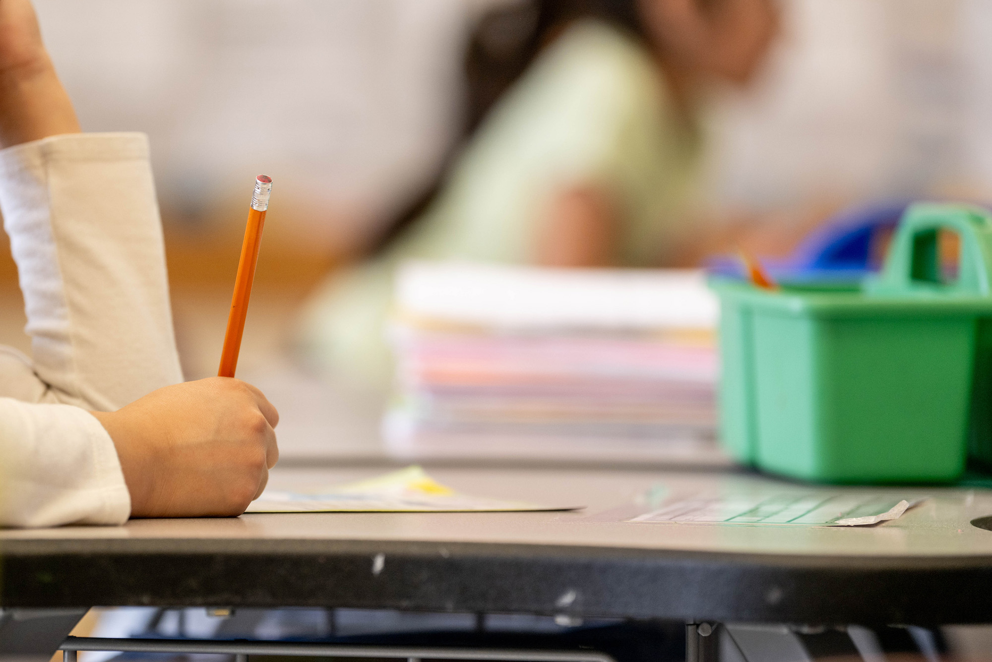 Photo: A close-up of an elementary student writing with a pencil with school supplies on their desk