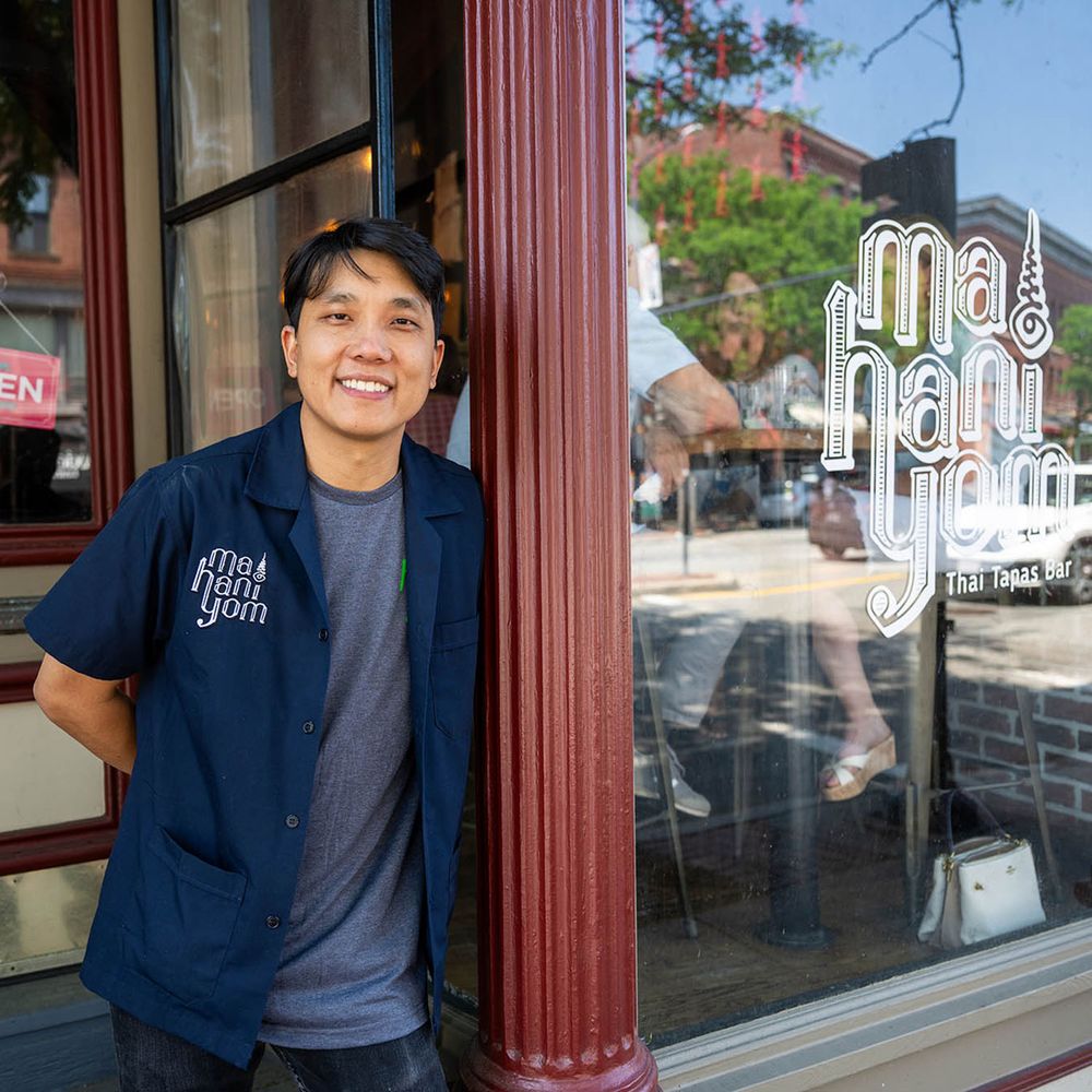 Photo: A young Asian man leans up against his store front. He is wearing a gray shirt and a navy button up.