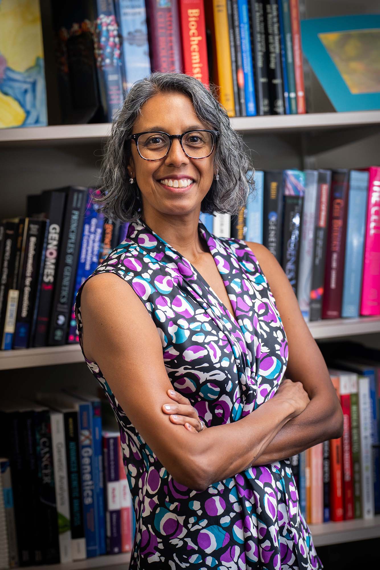 Photo: A portrait of a woman with chin length gray hair wearing a purple and blue sleeveless blouse. She smiles for the photo