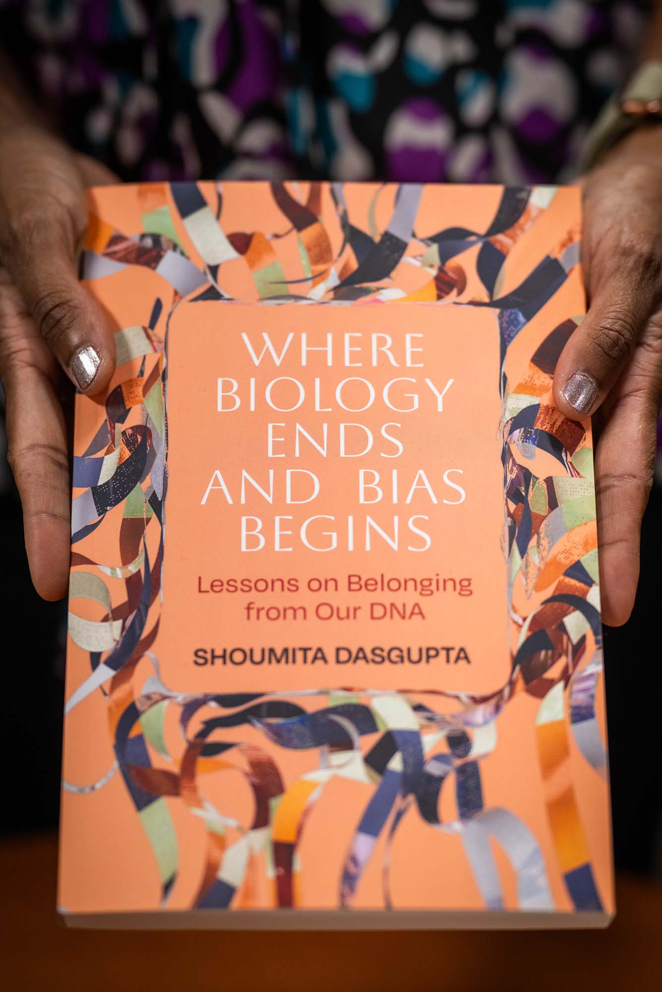 Photo: Two hands hold a soft orange colored book titled “Where Biology Ends and Bias Begins Lessons on Belonging from Our DNA