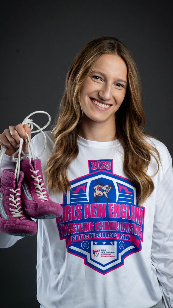 Photo: Hannah Bryson, a female wrestler at BU, holding up purple wrestling shoes and posing with a smile