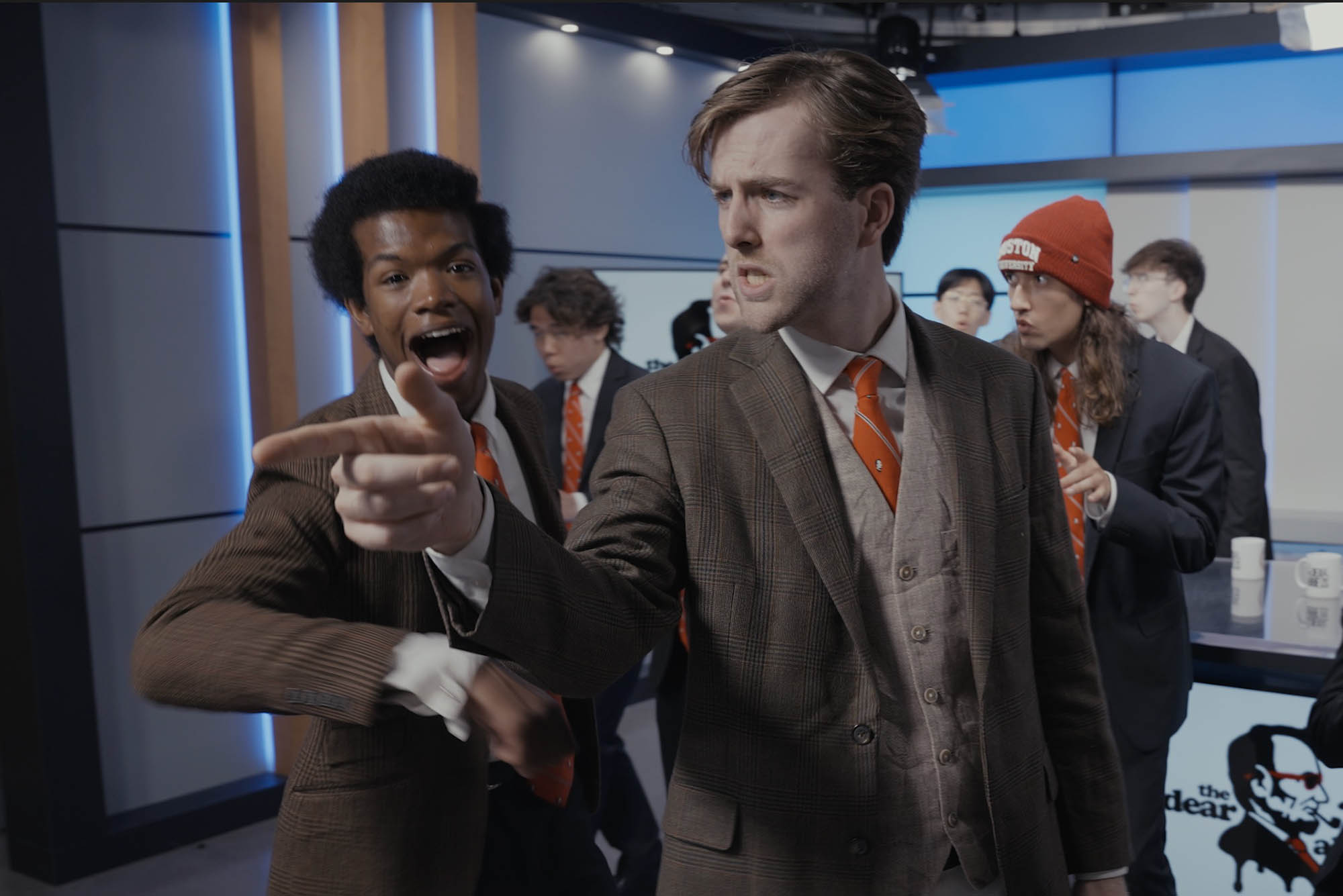 Photo: A still from a college production a cappella group in a newsroom. The students are dressed in suits.