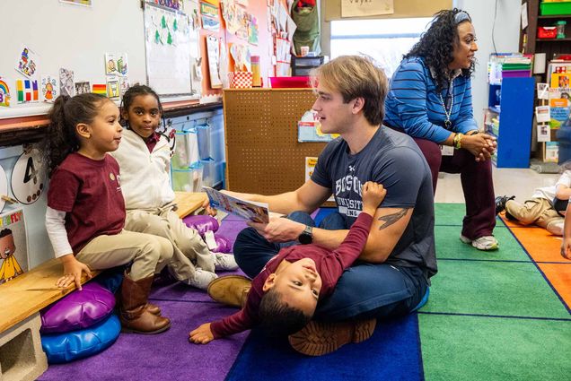 Photo: A young blonde man reads to small children for a BU event. A kid hangs off his lap