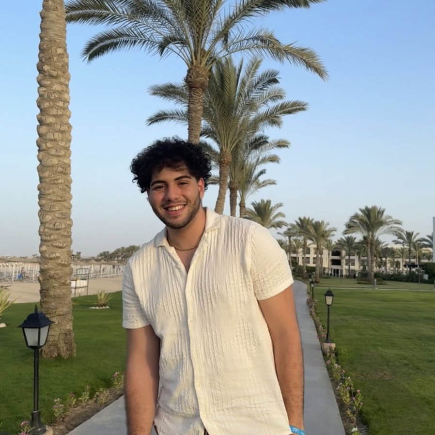 Photo: A person stands outdoors on a paved walkway lined with grass and palm trees, wearing a short-sleeved, light-colored shirt. Resort buildings and a clear blue sky are in the background.