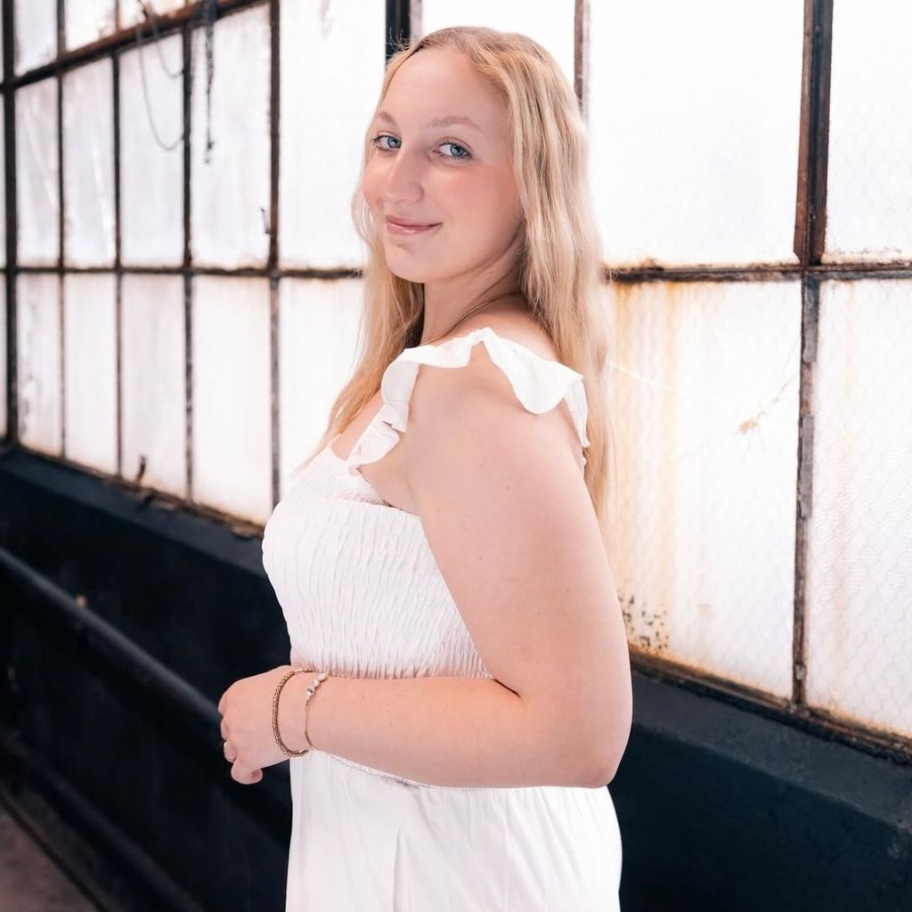 Photo: A person with long, light hair stands indoors near a wall of industrial-style windows wearing a white, ruffled, sleeveless dress, hands held together in front.